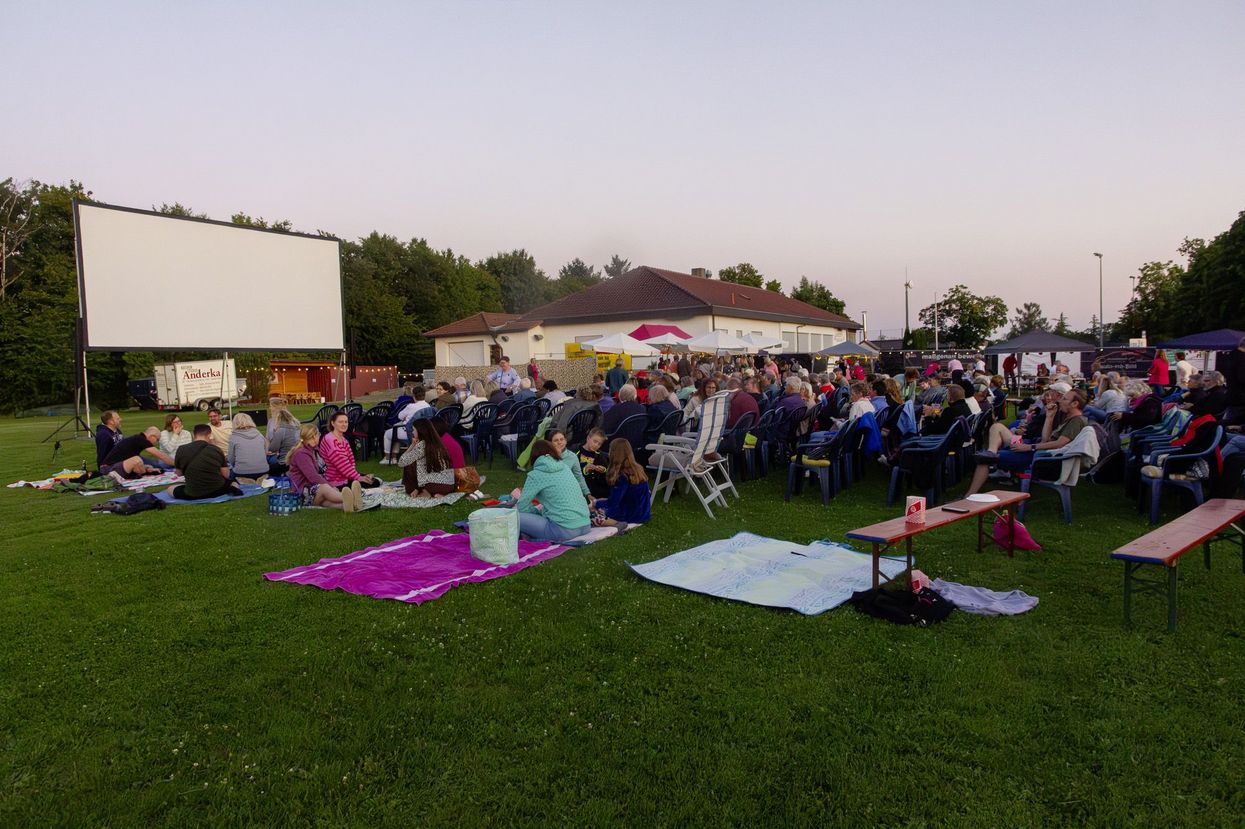 Links steht eine weiße Kinoleinwand. Im Hintergrund, in der Mitte, sieht man ein Gebäude. Auf der Wiese sitzen viele Menschen auf Stühlen. Im Vordergrund sitzen ca. 20 Menschen auf Picknickdecken auf der Wiese. Rechts im Vordergrund stehen zwei Bierbänke.