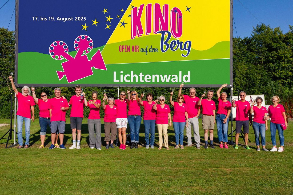 Mehrere Personen in einer Reihe vor einer Kinoleinwand auf einer Wiese. Auf der Leinwand ist das Plakat für das Open-Air-Kino in Lichtenwald zu sehen. Die Personen, die vor der Leinwand stehen, organisieren das Event. 