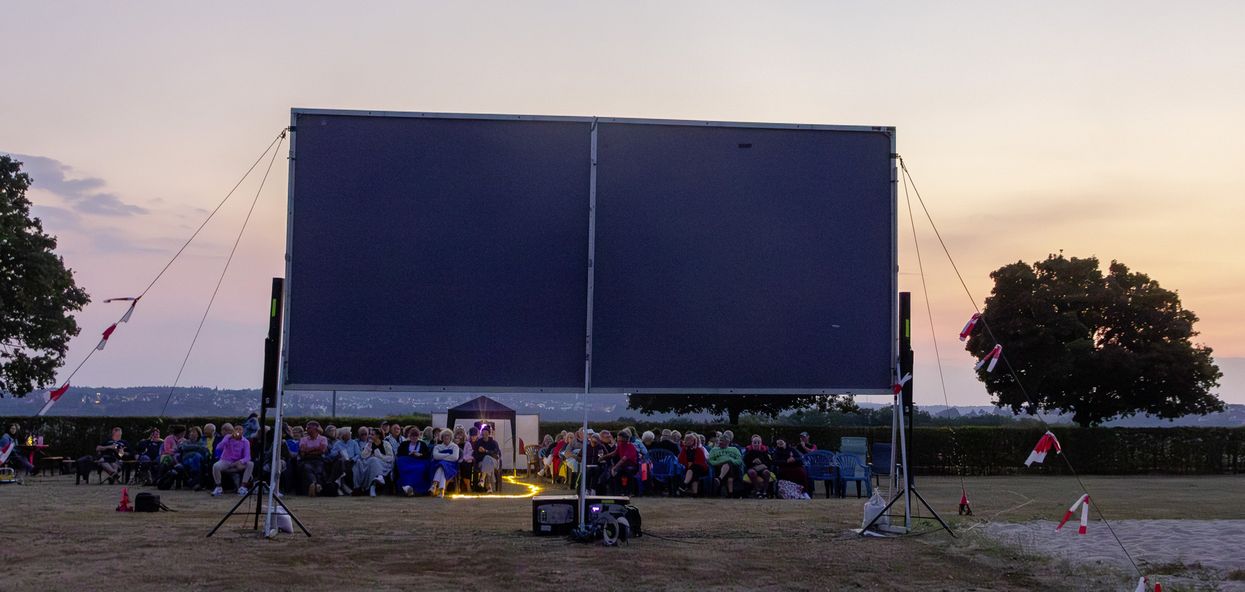Menschen sitzen beim Open-Air-Kino auf Stühlen vor einer Kinoleinwand. Im Hintergrund ist der Sonnenuntergang zu sehen.
