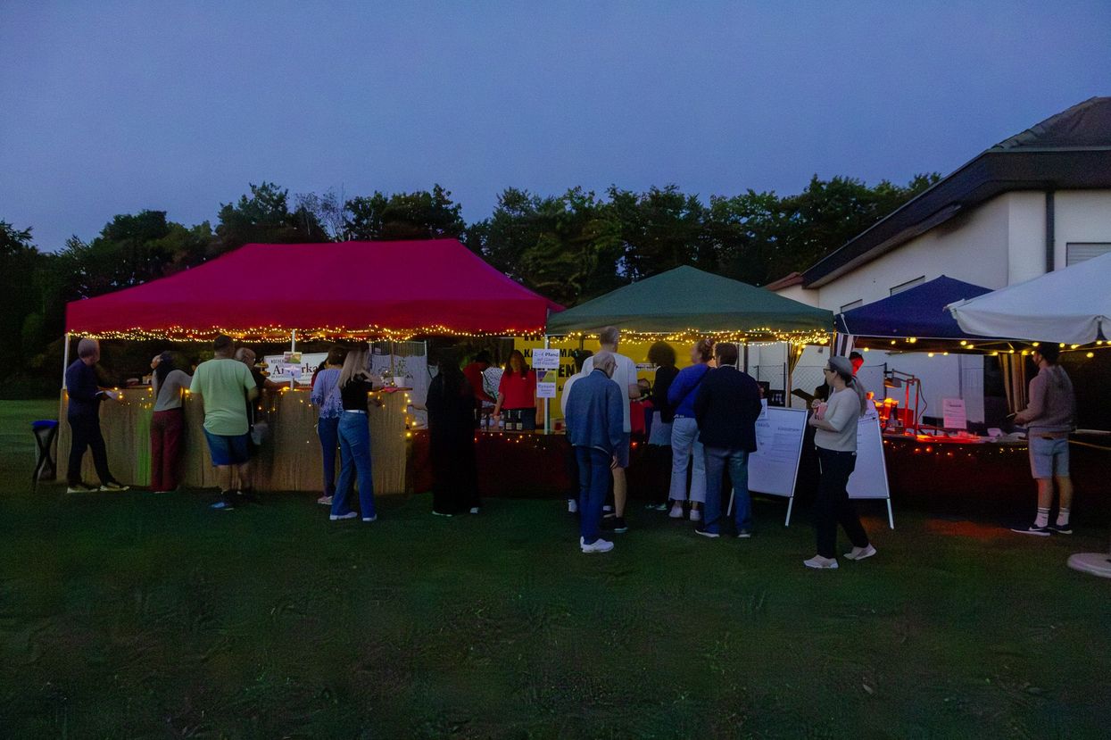 Es ist Abenddämmerung. Vor drei Pavillons, links ein roter, in der Mitte ein grüner und rechts ein blauer stehen, auf einer grünen Wiese, viele Menschen, um sich Getränke oder Essen zu holen. 